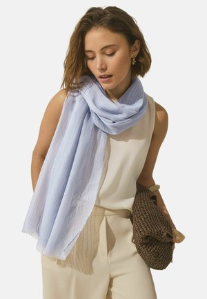 Kvinde med kort brunt hår bærer et lyseblåt tørklæde over en ærmeløs hvid top, beige bukser, og holder en brun flettet clutch.