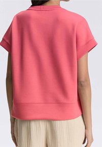 Koralkowy różowy sweatshirt z krótkim rękawem, okrągłym dekoltem i luźnym fasonem. Posiada ribbowany dół i miękką teksturę. Wyświetlony widok z tyłu.