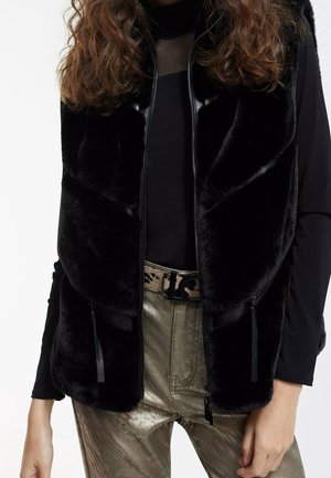 Vrouw die een zwart nepbonten gilet draagt over een zwarte top met lange mouwen, metalen goudkleurige broek en een riem met luipaardprint en een zwarte gesp.