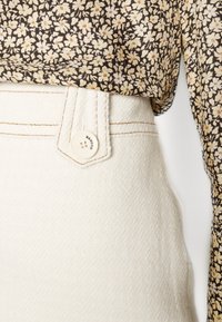 Białe, teksturowane spodnie o wysokim stanie z beżowymi szwami, z przodu zapinane na guzik. W zestawie z ciemnym topem w kwiatowy wzór.