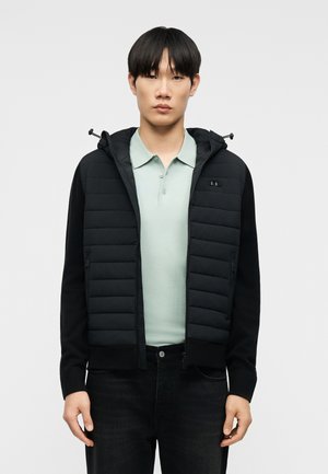 Noor mees kannab musta tikandiga lukuga jope kapuutsiga, selle all helerohelist polosärki, ta seisab ühevärvilise tausta ees.