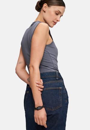 Graues ärmelloses Top mit Rundhalsausschnitt, kombiniert mit hochgeschnittenen, dunklen Jeans. Auffälliges Detail: schwarze Gliederkette am Handgelenk.