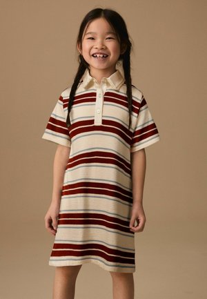 Lächelndes junges Mädchen mit geflochtenem Haar, das ein kurzärmeliges Kleid mit Kragen und horizontalen rot-weiß-blauen Streifen vor beigem Hintergrund trägt.