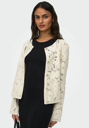 Kvinde iført sort kjole og cremefarvet hækleet cardigan, der holder en sort håndtaske mod en hvid baggrund.