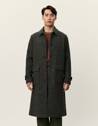 Σκούρο πράσινο μάλλινο παλτό σε σχέδιο ψαροκόκαλο με γιακά, μπροστινή κουμπότρυπα, δύο μεγάλες τσέπες με πατιλέτα και ρυθμιζόμενους καρπούς. Το μοντέλο στέκεται στραμμένο μπροστά.