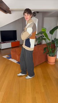 Pletený svetr s barevnými bloky v odstínech béžové, modré a oranžové; kombinovaný s volnými černými kalhotami a stříbrnými sportovními botami; dřevěná podlaha.