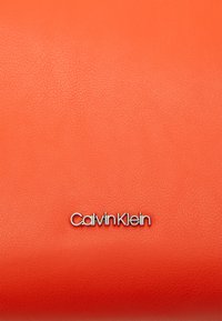 Narancssárga bőráru, amelynek textúrázott felülete van, és a fémes ezüst "Calvin Klein" logó jól láthatóan van elhelyezve.