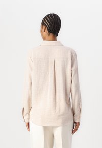 Chemise longue à manches beiges claires en tissu texturé, avec un col et un pli au dos. Présente des fentes sur les côtés des poignets pour un détail supplémentaire.
