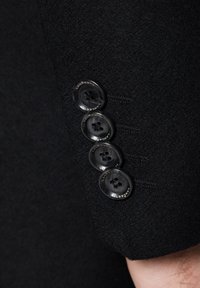 Rękaw czarnego wełnianego płaszcza z czterema okrągłymi, pasującymi guzikami z wygrawerowanym tekstem. Gładka faktura z drobnym detalem przeszycia wzdłuż krawędzi.