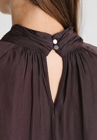 Mørk lilla bluse med høj hals og samlet stof, en nøglehulåbning bagpå og to sølvknapper til lukning.