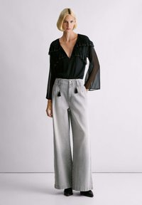 Kobieta z krótkimi blond włosami, ubrana w czarną przezroczystą bluzkę z długim rękawem i falbanami oraz w szerokie jasnoszare dżinsy, na jednolitym tle.