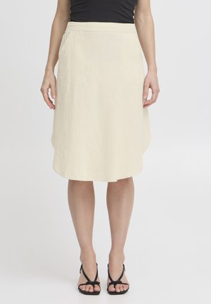 Žena stojaca na jednoduchom pozadí, oblečená v béžovej sukni po kolená, čiernom tielku bez rukávov a čiernych sandáloch na podpätku so šnurovaním a otvorenou špičkou.