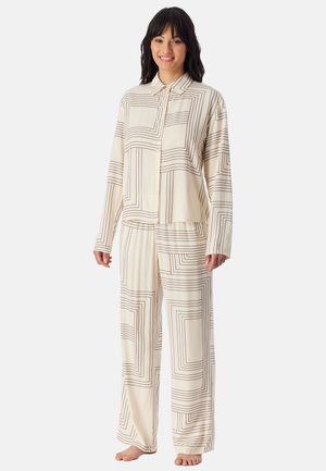 Smilende kvinde står barfodet iført kremfarvet løstsiddende pyjamas med sorte firkantede linjemønstre på top og bukser mod en ensfarvet baggrund.
