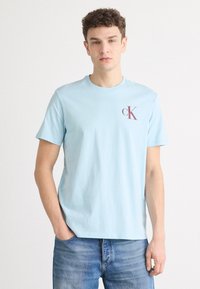 Světlemodré tričko s krátkým rukávem z bavlněného materiálu, s kulatým výstřihem, se tmavě vínovým logem "ck" na levé straně hrudi.