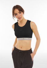 Donna che indossa un top corto sportivo nero con elastico bianco con la scritta "FREDDY" e pantaloni neri con coulisse, mani nelle tasche, su sfondo bianco.