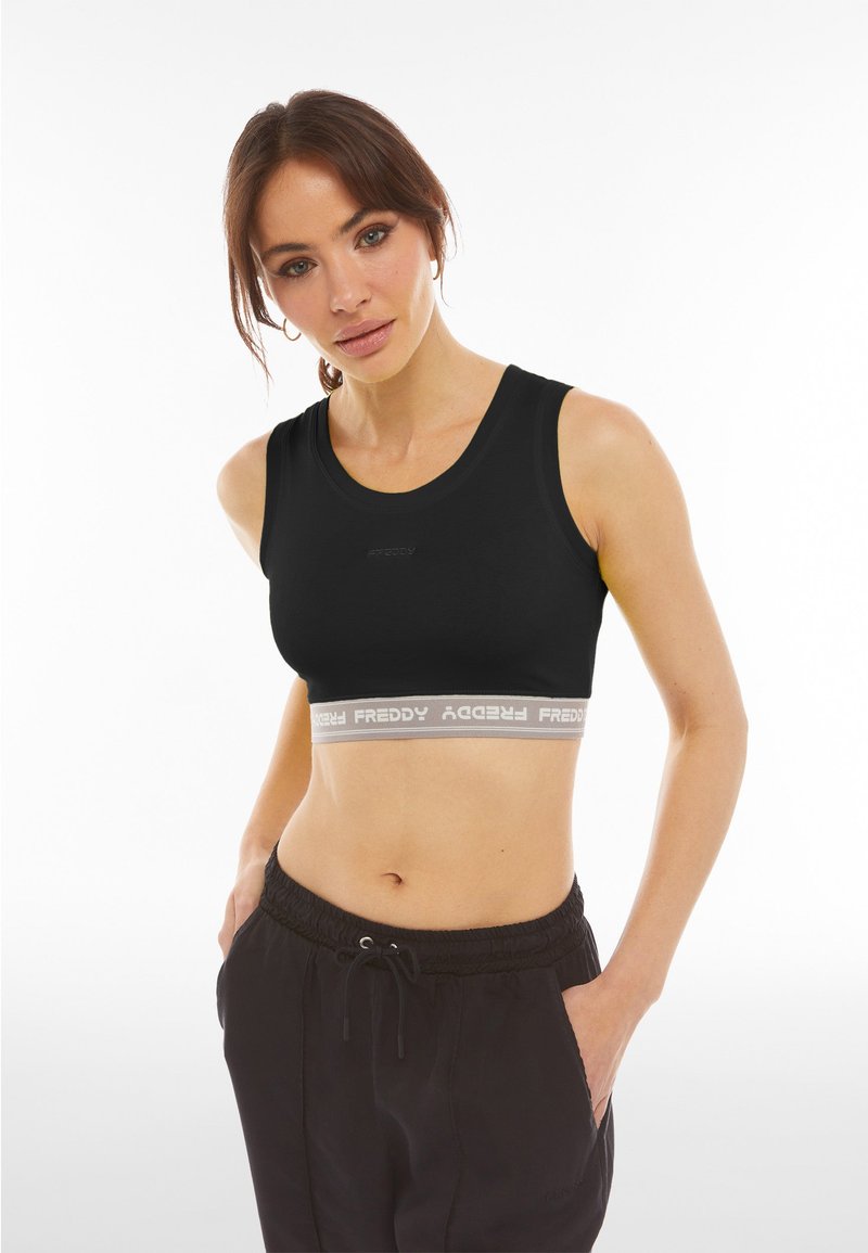 Donna che indossa un top corto sportivo nero con elastico bianco con la scritta "FREDDY" e pantaloni neri con coulisse, mani nelle tasche, su sfondo bianco.