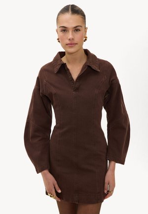 Vrouw die een nauwsluitende donkerbruine jurk met lange mouwen draagt, met wijde manchetten, een diepe kraag en gouden oorringen, tegen een effen witte achtergrond.