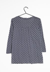 Blouse bleu marine à manches longues avec un motif géométrique blanc, col rond et détail froncé à l'avant. Suspendue à un crochet noir.