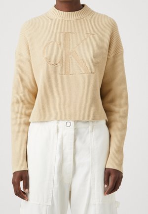 Osoba nosící béžový žebrovaný svetr s velkým logem "cK" a bílé kalhoty s vysokým pasem a zapínáním na kovový knoflík.