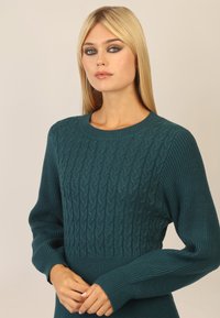Teal strikket sweater med et kabelstrikket mønster på forsiden og ribmede manchetter. Blødt materiale med lange ærmer og rund halsudskæring.