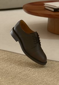 Tumši brūni ādas apavi ar saites, kas atrodas virs beigtas auduma paklāja, blakus apaļam koka galdam ar saliktām grāmatām.