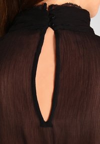 Blouse noire transparente avec un col haut en dentelle, un dos en keyhole, et deux boutons noirs au niveau du cou, créant un aspect texturé.