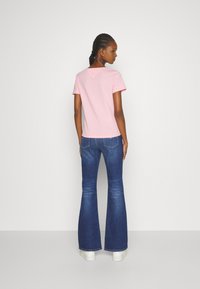 Camiseta rosa de manga corta con un detalle en forma de V en la parte posterior, combinada con unos vaqueros oscuros de pernera acampanada y zapatillas blancas. Diseño y colores sencillos.
