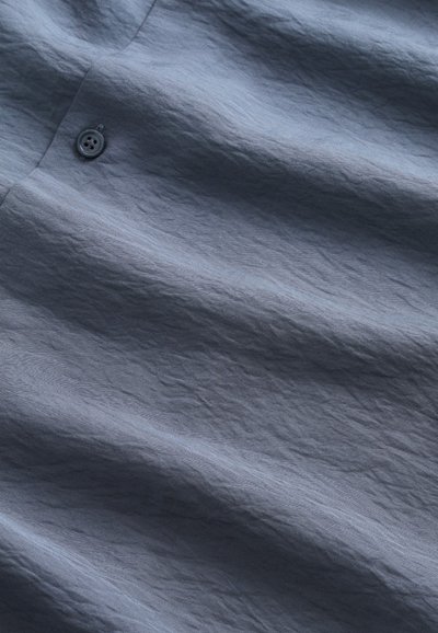 Tissu texturé dans une couleur bleu-gris profond, présentant des plis visibles. Un détail de bouton unique est présent, améliorant le design.