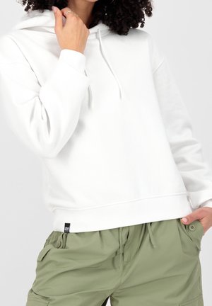 Femme portant un sweat à capuche blanc avec cordons et un pantalon cargo vert clair, une main près du visage et une main dans la poche.