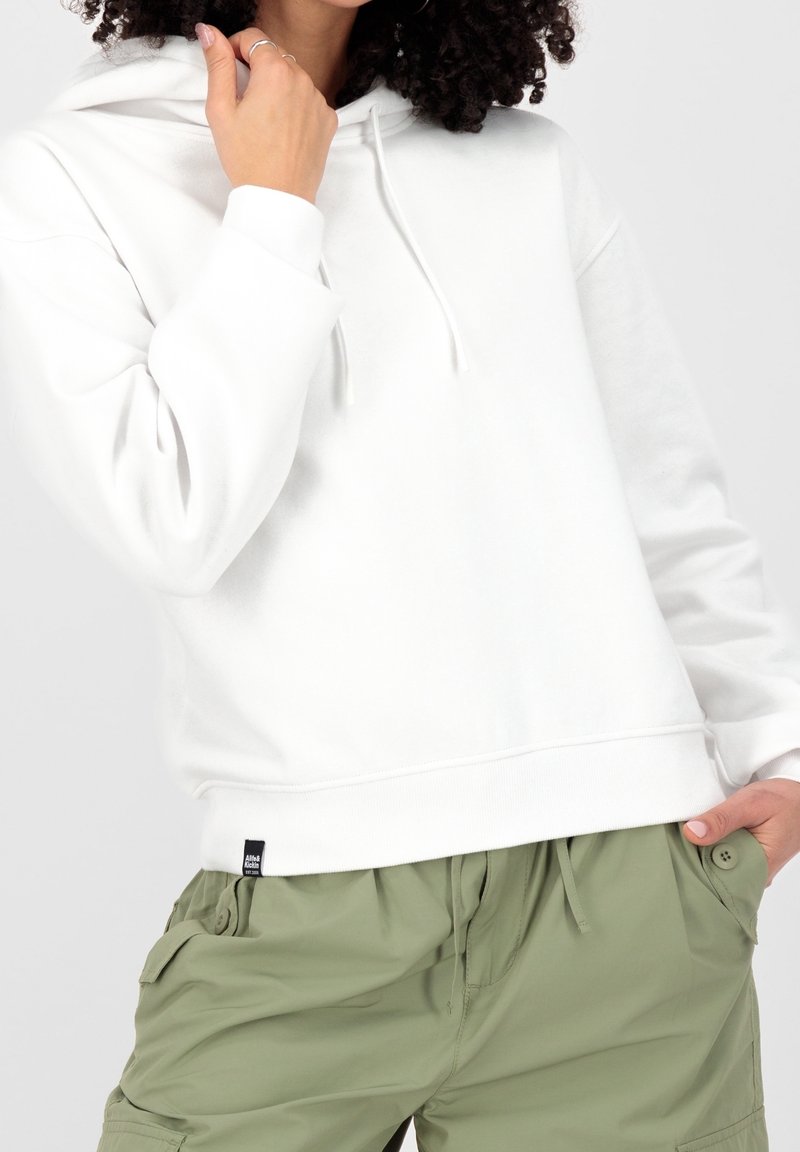 Femme portant un sweat à capuche blanc avec cordons et un pantalon cargo vert clair, une main près du visage et une main dans la poche.