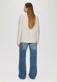 Kobieta z długimi prostymi włosami, ubrana w biały sweter z ciemnymi paskami na rękawach i luźne niebieskie dżinsy, stojąca tyłem.