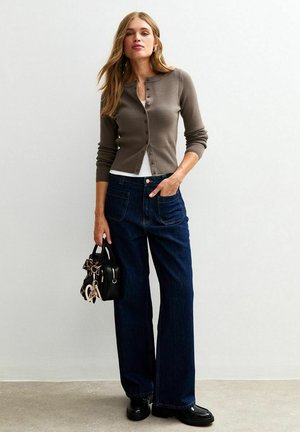 Bruine gebreide cardigan over een witte blouse, gecombineerd met losse blauwe denim jeans. Accessoires zijn onder andere een kleine zwarte handtas met een charm.
