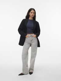 Vrouw draagt een zwart colbert, een marineblauwe cropped top, lichtgrijze high-waisted jeans en zwarte puntige schoenen, staand met handen in de zakken.