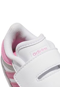 Baskets blanches avec des accents roses et des sangles élastiques. Tige en synthétique lisse, semelle en caoutchouc et doublure en mesh pour la respirabilité. Logo distinct présent.