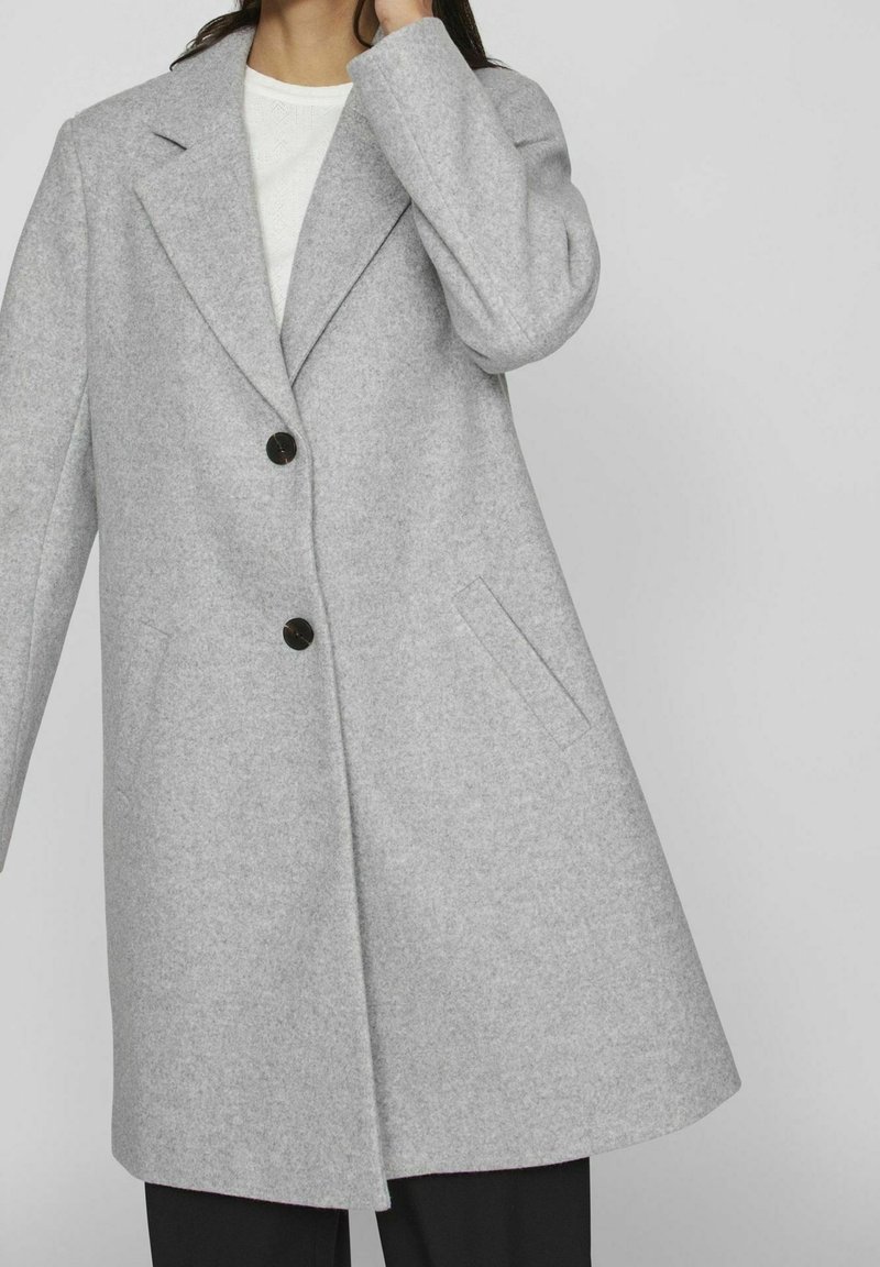 Femme portant un manteau en laine gris clair avec deux boutons noirs, de larges revers et des poches latérales par-dessus un haut blanc.