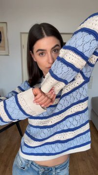 Νεαρό άτομο με ίσια σκούρα μαλλιά, που φοράει πλεκτό πουλόβερ με μπλε και λευκές ρίγες και μπλε τζιν, ποζάρει με τα χέρια σταυρωμένα σε ένα δωμάτιο.