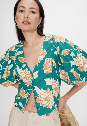 Femme portant un chemisier à boutons bleu sarcelle à motifs floraux avec des manches bouffantes, un pantalon beige, des boucles d'oreilles en argent en forme de créoles, et tenant un sac en paille tressée.