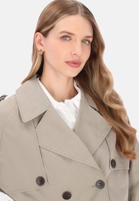 Beiger Trenchcoat mit großen Revers, doppelt geknöpften braunen Knöpfen und glatter Textur, kombiniert mit einem weißen Hemd mit Kragen darunter.