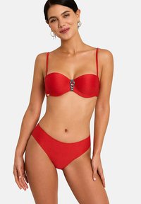 Femme portant un bikini deux pièces rouge texturé avec une boucle décorative sur le haut et des bretelles fines, posant avec un léger sourire.