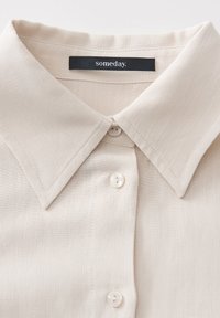 Beige blouse with a large point collar, a button-down front, and pearl-like buttons; made of smooth, lightweight fabric with subtle texture.