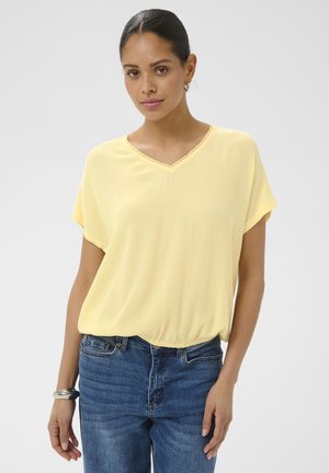 Kobieta ubrana w jasnożółtą bluzkę z krótkim rękawem i dekoltem w serek oraz niebieskie dżinsy, pozująca z rękami swobodnie opuszczonymi wzdłuż ciała na jednolitym tle.