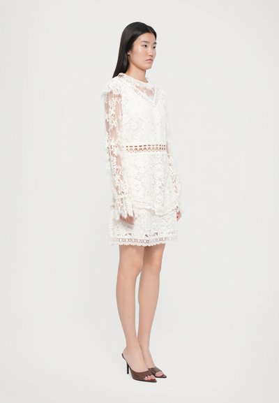 Λευκό δαντελένιο φόρεμα με μακριά διάφανα μανίκια, ντεκολτέ σε σχήμα V και κοψίματα στη μέση και το στρίφωμα. Φορεμένο με καφέ ανοιχτά τακούνια.