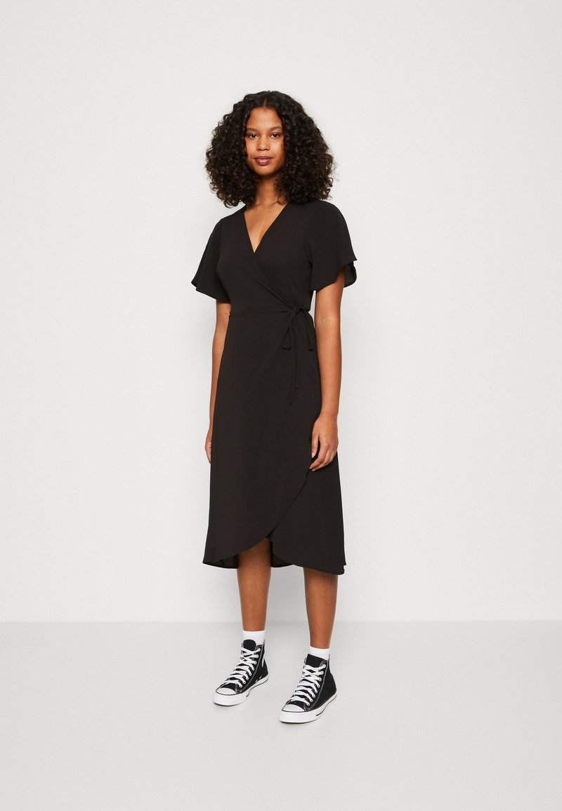 Robe noire à manches courtes avec un lien à la taille, associée à des baskets montantes noires et des chaussettes blanches mi-mollets.