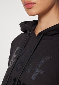 Gros plan d'une personne portant un sweat à capuche noir avec le texte scintillant "DKNY SPORT NYC 1989" et des cordons noirs, montrant le bas du visage et le cou.