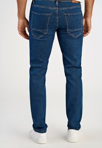 Ciemnoniebieskie dżinsy, slim fit, z dwiema tylne kieszeniami ozdobionymi przeszyciami, noszone z białymi sneakersami. Gładka tekstura, minimalistyczny design.