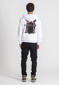 Biela mikina s kapucňou so grafikou troch psov na chrbte, s textom "KAVANAGY." Skombinovaná s čiernymi cargo nohavicami a vzorovanými teniskami.