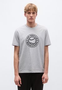 Pilkas medvilninis marškinėlis su apvaliu kaklu, trumpomis rankovėmis ir priekyje esančiu juodu apskritimo spaudiniu, vaizduojančiu teniso temą ir prekės pavadinimą.