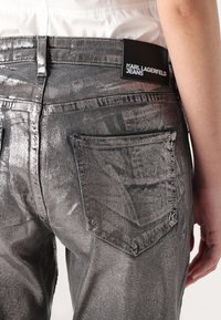 Pantalones vaqueros metálicos plateados con corte ajustado, bolsillos traseros y una etiqueta negra con el logotipo "KARL LAGERFELD JEANS". Acabado texturizado.