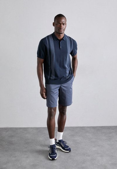 Marineblaues Poloshirt mit vertikalen Streifen, kombiniert mit hellgrauen Shorts. Das Modell trägt dunkle Sneakers, weiße Socken und steht auf einem grauen Boden.