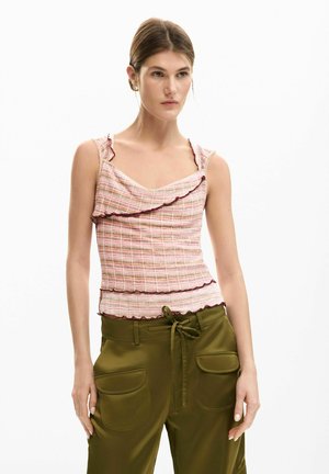 Jeune femme portant un haut sans manches rose à volants et un pantalon vert olive avec de grandes poches, debout devant un fond blanc.